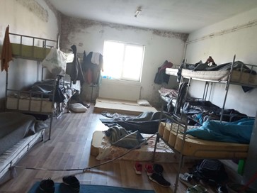Color image of a dorm at the refugee center. On the two sides there are two bunk beds, with several more mattresses laid on the ground in between. The walls are brown from dirt or mold in many places and there are clothes and shoes scattered around the room. Photo sent to Dreptul la Oraș